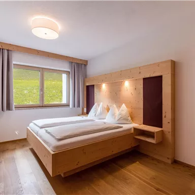 A modern bedroom with a wooden double bed. Large windows let in plenty of daylight and offer a view of the greenery.