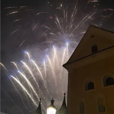 Ein wunderschönes Feuerwerk erleuchtet den Nachthimmel, hinter einem historischen Gebäude. Die Glühbirne einer Laterne strahlt im Vordergrund.