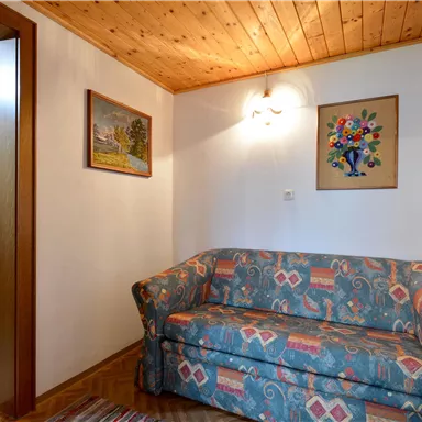 A cozy living room with a colorful sofa and wooden floor. Two pictures hang on the wall, and a bathroom is visible in the background.