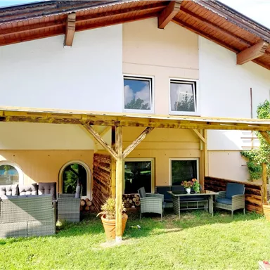 Ein gemütliches Haus mit einer überdachten Terrasse und einer Gartenmöbelgarnitur. Der Rasen ist grün und es gibt einige Pflanzen in Töpfen.