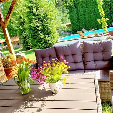 Ein gemütlicher Gartenbereich mit einem Tisch und bequemen Sitzkissen. Im Hintergrund ist ein Pool und üppige grüne Pflanzen zu sehen.