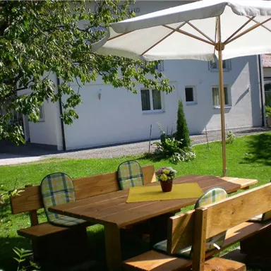 A cozy wooden garden table with benches and a sun umbrella. Surrounded by green spaces and plants in a quiet garden.