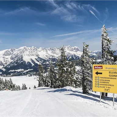 A snow-covered mountain landscape with tall fir trees and majestic peaks. A sign points the way to the ski slopes to Ellmau and Going.