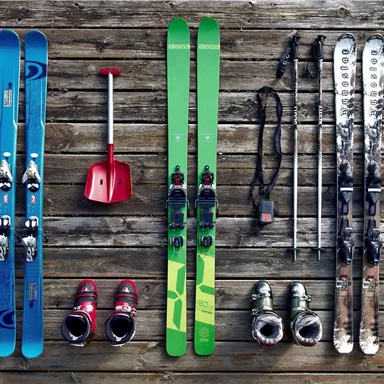 Two pairs of skis with matching ski boots are lying on a wooden floor. A shovel and two ski poles are also visible.