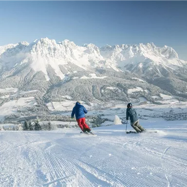 Two skiers on a snowy slope with majestic mountains in the background. The sky is clear, and the landscape is wintery beautiful.