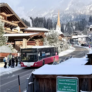 Ein Winterblick auf ein malerisches Dorf mit schneebedeckten Bergen. Ein Bus hält an einer Haltestelle, während Passagiere darauf warten.
