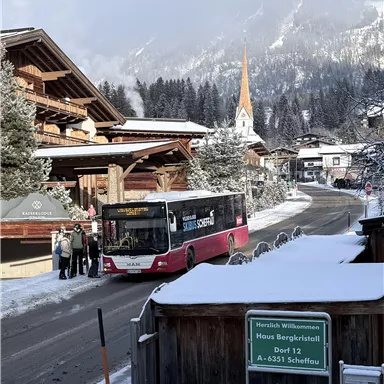 A bus stops in a snowy village. The background shows mountains and a church with a pointed tower.