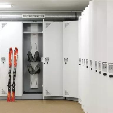 A modern ski room with white lockers. Two skis and one set of ski equipment are stored in one of the lockers.