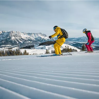Two skiers are skiing on a snow-covered slope. In the background, impressive mountains and a clear sky can be seen.
