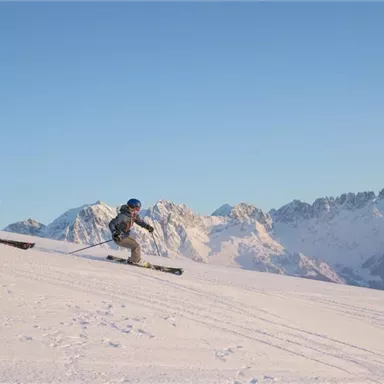 Two skiers are riding on a snow-covered slope. In the background, impressive mountains and a blue sky are visible.