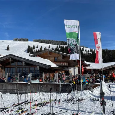 A ski lodge with many people standing in the snow. Surrounded by mountains and ski lifts, flags are blowing in the wind.