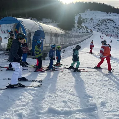 A group of children is standing in a ski line on a snow-covered slope. In the background, a conveyor belt and forests can be seen.