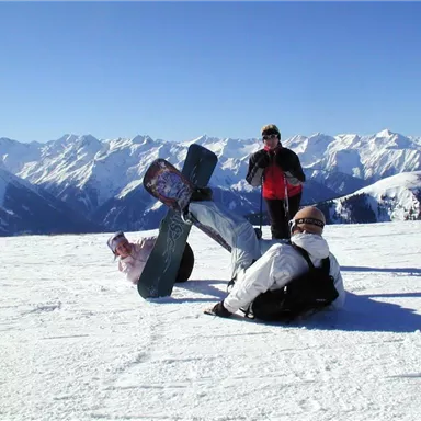 Drei Personen stehen auf einem schneebedeckten Berggipfel. Im Hintergrund sind hohe Berge und blauer Himmel zu sehen.