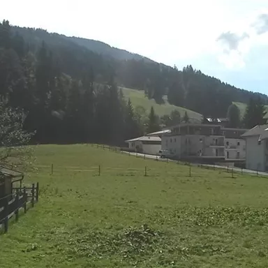 A picturesque landscape with green meadows and hilly forests. In the background, there are several buildings that are harmoniously integrated into nature.