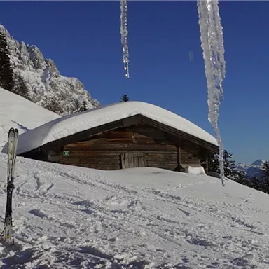 Eine gemütliche Hütte im Schnee mit Skiern im Vordergrund. Der Himmel ist klar und die Berge sind im Hintergrund sichtbar.