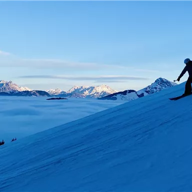 A skier descends a snowy slope. In the background, there are mountains and a bright blue sky.