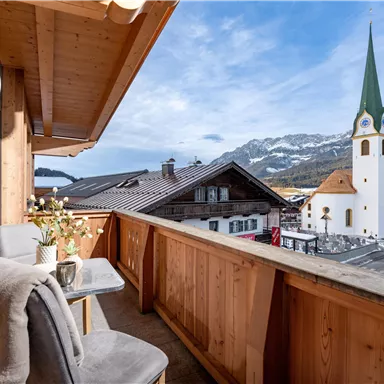 A beautiful balcony with a view of the mountains and a church. The surrounding wooden houses and the clear blue sky create a cozy atmosphere.