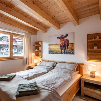 A cozy bedroom with a double bed and wooden furniture. A picture of a cow hangs on the wall, and a window lets in plenty of daylight.