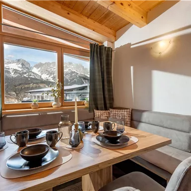 A cozy dining area with a wooden table and dishes. In the background, the mountains can be seen through the large window.