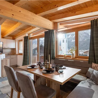 A cozy dining area with wooden furniture and large windows. The view of the mountains and the daylight create a warm atmosphere.