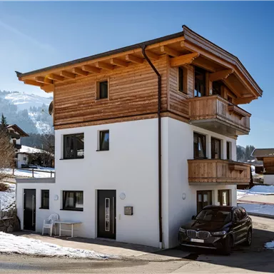 A modern house with a wooden chair and a car in front of the door. The surroundings are covered in snow and the mountains are visible in the background.