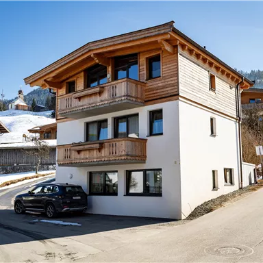 A modern, two-story house with wooden balustrades in a snow-covered landscape. In the foreground, there is a car, and in the background, mountains can be seen.