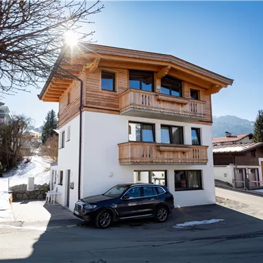 A modern house with wooden elements and large windows. Snow lies on the ground and the mountains are visible in the background.