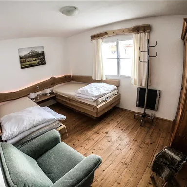 Ein einfaches Zimmer mit zwei Betten und einem gemütlichen Sessel. Der Holzfußboden und die hellen Fenster schaffen eine einladende Atmosphäre.