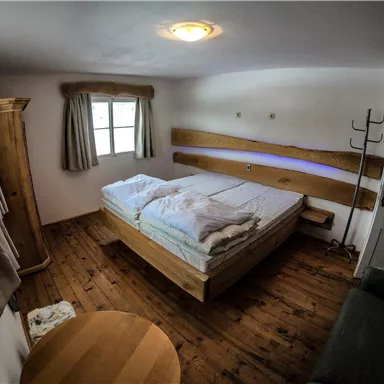 Ein einfaches Schlafzimmer mit einem Doppelbett und Holzmöbeln. Das Zimmer hat ein Fenster mit Vorhängen und einen kleinen Tisch.
