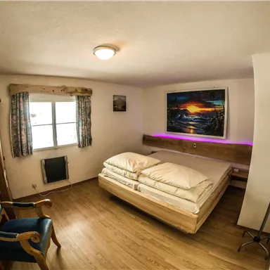 Ein gemütliches Schlafzimmer mit einem Doppelbett und einem blauen Sessel. An der Wand hängt ein farbenfrohes Bild eines Sonnenuntergangs.