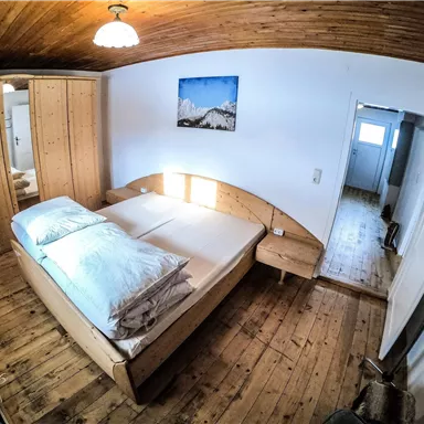 Ein gemütliches Schlafzimmer mit einem Holzbett und großen Kissen. Die Wände sind hell gestrichen und die Holzdecke gibt einem warmen Eindruck.