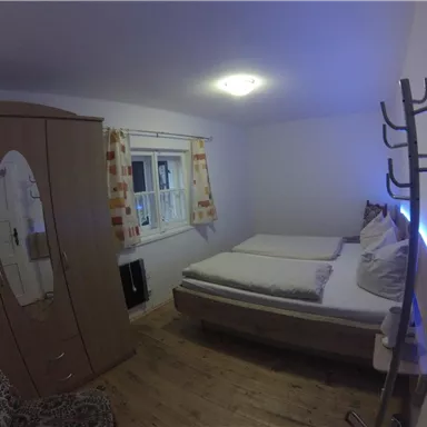 Ein einfaches Schlafzimmer mit einem Doppelbett und einem Kleiderschrank. Die Wände sind hell, und es gibt ein Fenster mit Vorhängen.