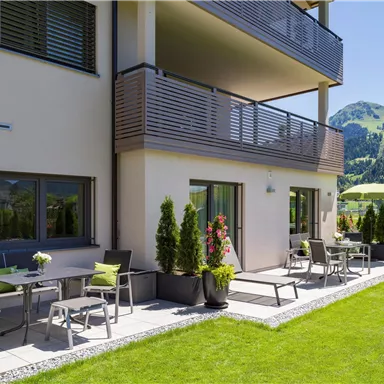 A modern terrace with seating and a well-kept garden. In the background, green mountains and a blue sky are visible.