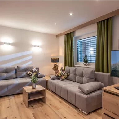 A modern living room with gray sofas and a wooden table. The room has large windows and is decorated with plants.