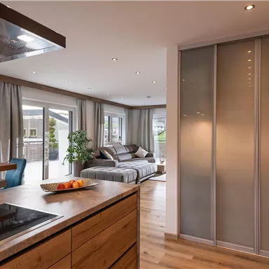 A modern apartment with an open living area and a wooden dining table. The room is bright and stylishly furnished with large windows and a gray sofa set.