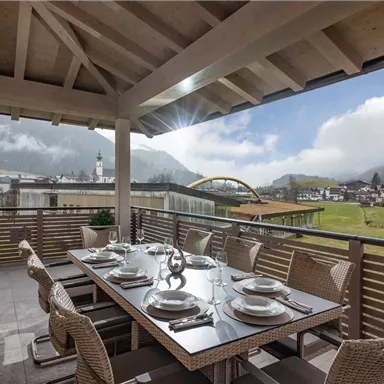 A beautiful dining area on a terrace overlooking a picturesque landscape and clouds. The table is set with dishes, and the surrounding mountains are visible.