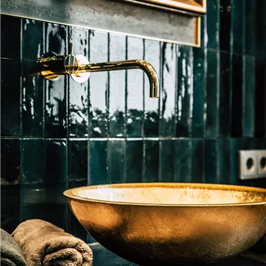 An elegant bathroom with a golden washbasin and a modern faucet. In the background, dark tiles and a golden mirror can be seen.