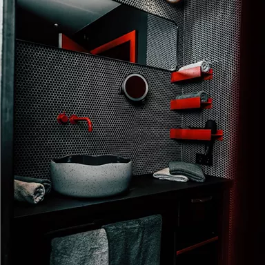 A modern bathroom with a gray washbasin and red accents. The walls are designed with a unique pattern, and there are towels and shelves in matching colors.