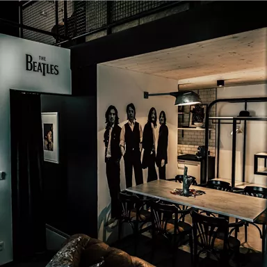 A room with a table and chairs, decorated with pictures and a large wall graphic. The wall features the silhouettes of a famous music band.