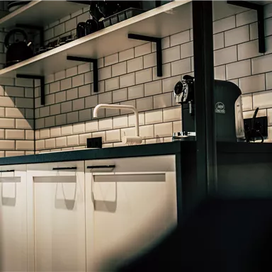A modern kitchen with white cabinets and a black countertop. On the wall, there are white tiles and a coffee machine visible.