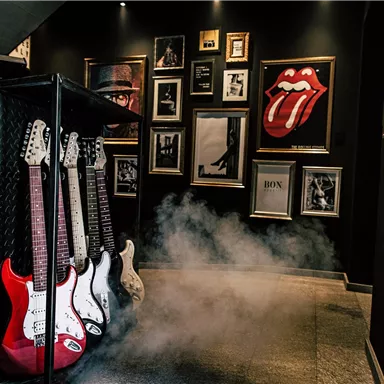 A stylish room with a wall full of music posters and frames. In the foreground are several electric guitars, while the floor is covered with light mist.