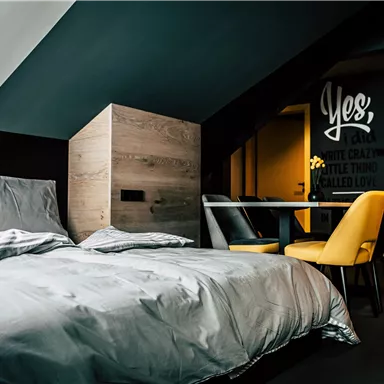 A modern bedroom with a comfortable bed and stylish furniture. The walls are painted in dark colors, and a table with yellow chairs complements the room.