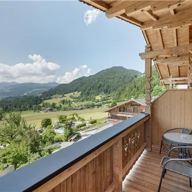 Ein Balkon mit Blick auf eine grüne Landschaft und Berge. Auf dem Balkon stehen zwei Stühle und ein kleiner Tisch.