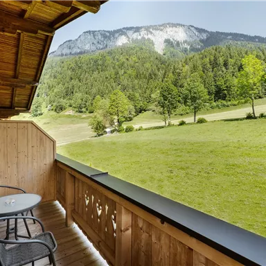 Ein Balkon mit zwei Stühlen und einem Tisch, der einen Blick auf eine grüne Wiese und Berge im Hintergrund bietet. Die Umgebung ist ruhig und umgeben von Bäumen.