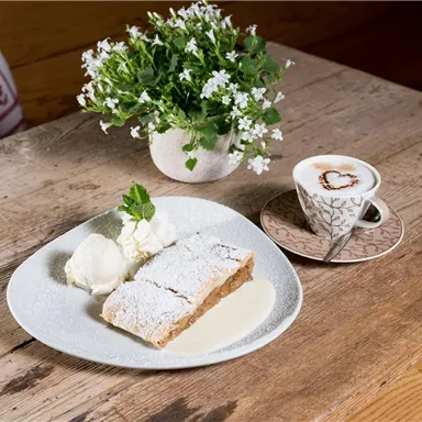 A plate with apple strudel, cream, and vanilla sauce is on a wooden table. Next to it is a cup of coffee with a heart design and a small pot of flowers.
