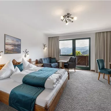 A modern hotel room with a large bed, a table, and chairs. The view from the window shows a beautiful mountain landscape.