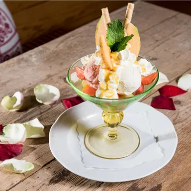 A seductive ice cream sundae with various flavors of ice cream, cream, and fresh fruits. Surrounded by rose petals on a wooden table.