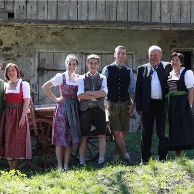 A group of six people in traditional attire stands in front of an old farmhouse. The surroundings are green and sunny.
