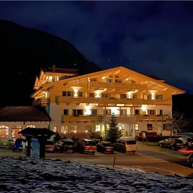 A cozy hotel at night, illuminated by warm light. In the foreground are some cars and snow-covered ground.