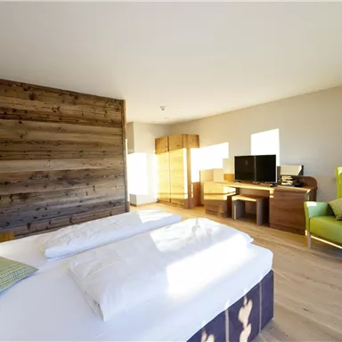 A modern bedroom with a large bed and a wooden panel. The room is bright and minimally furnished, with a cozy armchair and a desk.